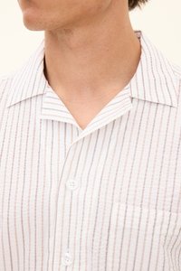 Homme portant une chemise blanche boutonnée avec de fines rayures verticales marron et un col ouvert, laissant apparaître une partie de son cou et de son menton.