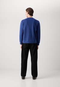 Suéter azul de mezcla de algodón, con un cuello y puños acanalados. Llevado con pantalones sueltos negros, vista desde atrás.