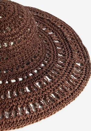 Hnědý široký klobouk z pletené slaměné slámy s průhledným vzorem a texturovaným copánkovým designem, vystavený na bílém pozadí.