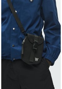 Sac bandoulière en tissu noir avec fermeture à rabat, sangle réglable et détail logo. Porté sur une veste bleue boutonnée et un pantalon noir.