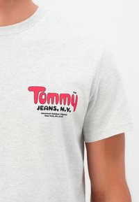 Gros plan sur une personne portant un T-shirt gris clair avec le texte rouge et noir "Tommy Jeans, N.Y." imprimé sur le côté gauche de la poitrine.