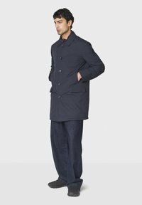 Dunkelblauer Steppmantel mit hohem Kragen, Knopfverschluss und zwei Seitentaschen. Trägt eine lockere dunkle Jeans und schwarze Sportschuhe.