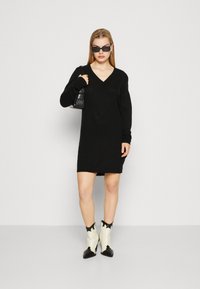 Vestido preto de malha com decote em V e mangas longas, combinado com botas estilo cowboy brancas e pretas e uma pequena bolsa preta.