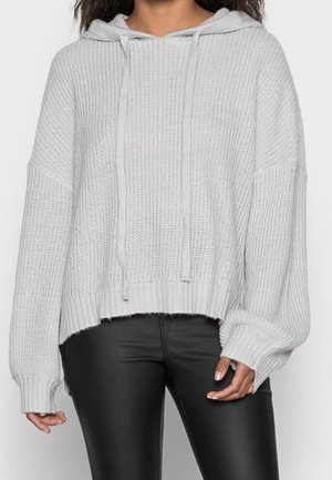 Femme portant un sweat à capuche en maille gris clair avec cordons de serrage et un pantalon en similicuir noir, mains détendues le long du corps.