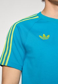 T-shirt bleu à manches courtes avec des accents jaune vif à trois bandes sur les épaules et un logo brodé sur la poitrine. Texture lisse.