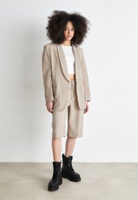 Blazer di lino beige abbinato a pantaloni corti beige a lunghezza ginocchio, indossato sopra un top corto bianco. Stivali neri con lacci spessi completano l'outfit.