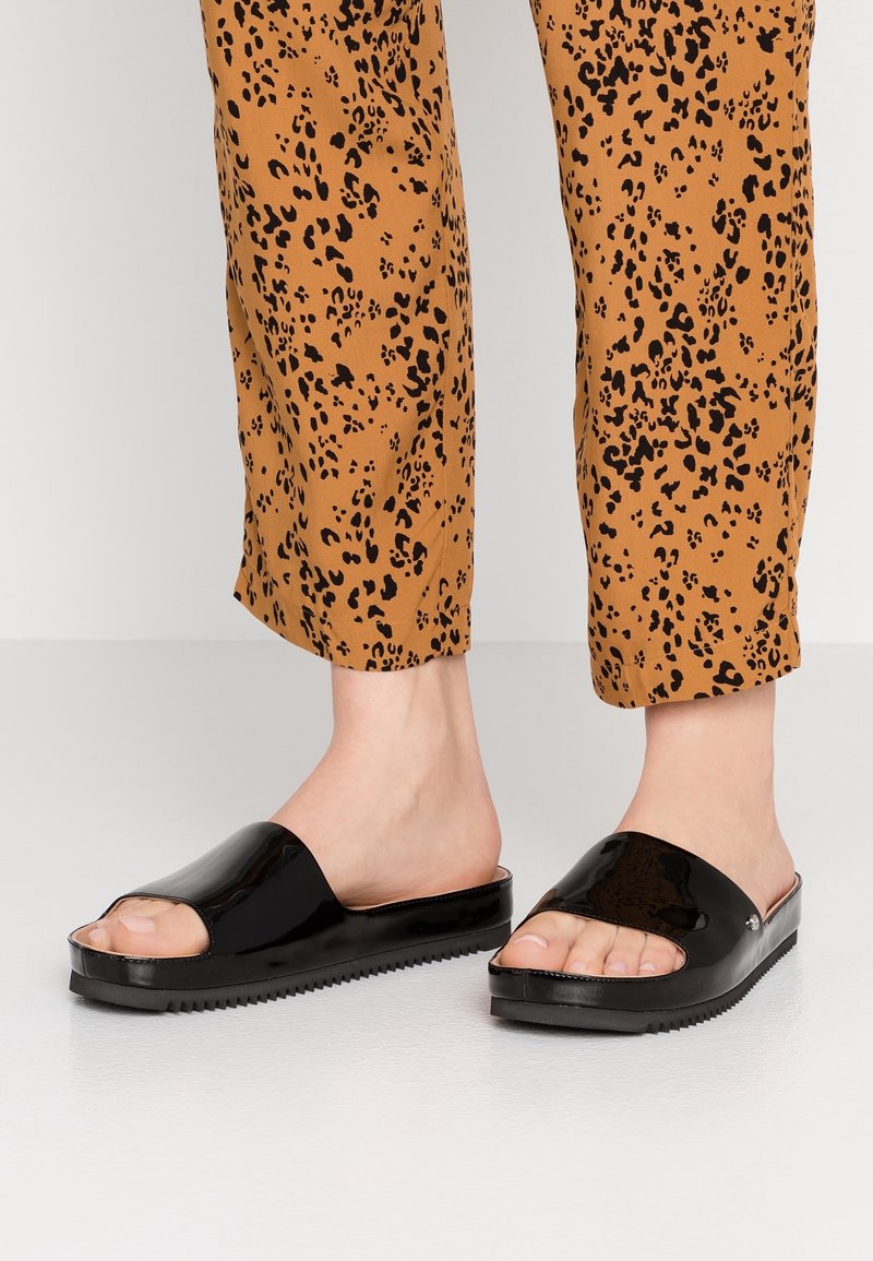 Jambes portant un pantalon marron à motif léopard et des sandales noires ouvertes à l'avant avec des semelles texturées sur un sol clair.