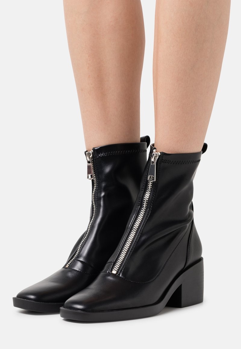 Paire de bottines en cuir noir avec fermetures éclair argentées à l'avant et talons blocs portées sur des jambes nues sur fond blanc.