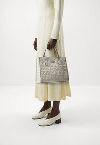 Silberne gesteppte Handtasche mit doppelten oberen Griffen, die ein elegantes Design und ein dezentes Logo-Detail aufweist, kombiniert mit einem hellgelben Kleid und weißen Schuhen.