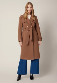 Brąszawe wełniane trencz, z podwójnymi guzikami, w talii z paskiem i zrolowanymi mankietami, noszone na niebieskich szerokich dżinsowych spodniach.