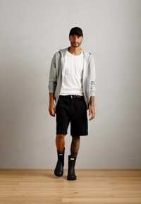 Homme marchant à l'intérieur portant un sweat à capuche gris, un t-shirt blanc, un short noir, une casquette noire et des bottes en caoutchouc noires sur un sol en bois.