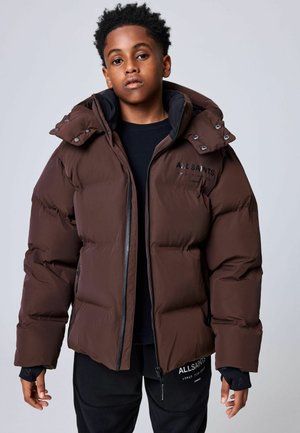Jong persoon draagt een bruine donzen jas en zwarte kleding, staat voor een effen lichte achtergrond, kijkt recht vooruit met een neutrale gezichtsuitdrukking.