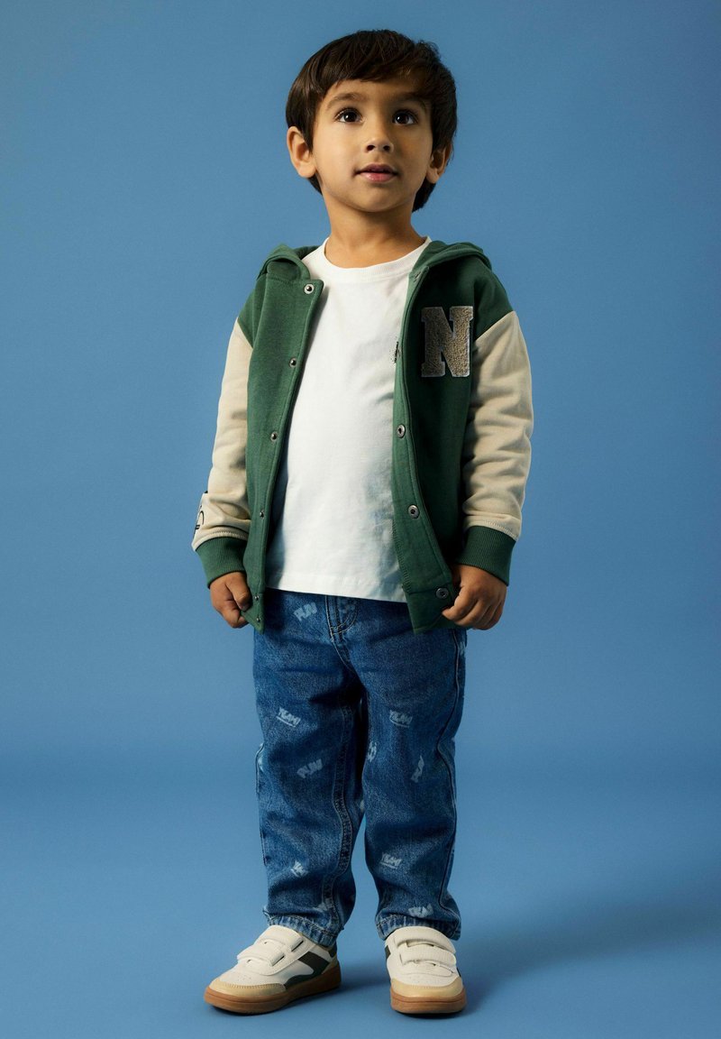 Sudadera verde con mangas beige, camiseta blanca y jeans azules con ligero desgaste. Zapatillas blancas con suelas marrones y detalles grises.