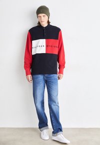 Long-sleeve polo shirt in navy, red, and white with branded text; paired with blue jeans and white sneakers; olive beanie on head.