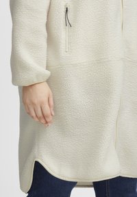 Krämfärgad fleecejacka med mjuk textur, med en zip-ficka, rundad nederkant och kontrasterande kant. Hand som vilar på tyget.