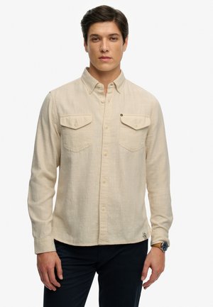 Jeune homme portant une chemise beige à boutons avec poches poitrine et un pantalon foncé, debout devant un fond blanc uni.