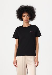 Black cotton t-shirt with short sleeves, featuring a small red "GUESS" logo on the left chest. Fabric appears smooth with a relaxed fit.