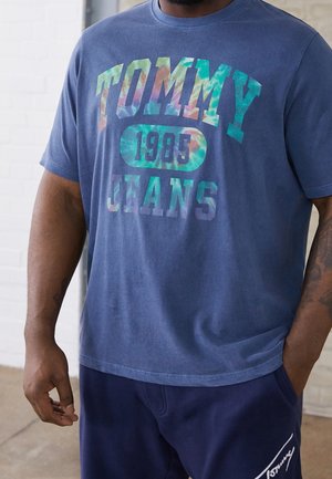 Hombre con una camiseta azul tie-dye Tommy Jeans 1985 y pantalones cortos azul marino con el logo de Tommy en la pierna, de pie en interiores.