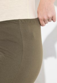 Olivgröna leggings gjorda av mjukt tyg, med resårmidja och slät textur, hålls upp i midjan.