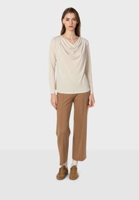 Beige Oberteil mit Kapuze und langen Ärmeln kombiniert mit braunen weiten Hosen. Das Model trägt braune Sneakers und präsentiert eine weiche, glatte Stofftextur.