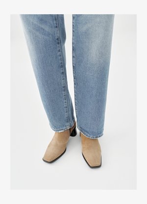 Beige suède enkellaarsjes met een puntige neus en een lage zwarte blokhak, gedragen met lichtblauwe jeans met rechte pijpen.