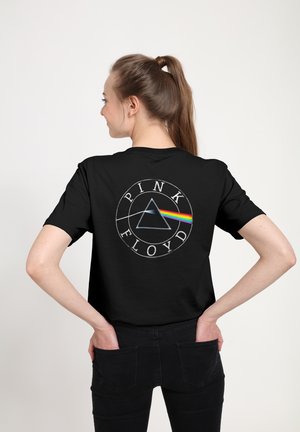 Kobieta z brązowym kucykiem, ubrana w czarną koszulkę Pink Floyd z grafiką pryzmatu i tęczy, ręce w tylnych kieszeniach, stojąca na jednolitym tle.