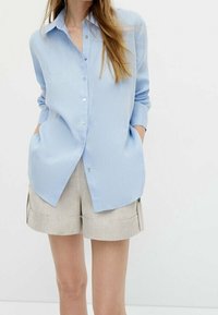 Hellblaues Button-Down-Shirt mit spitzem Kragen, langen Ärmeln und zwei seitlichen Taschen, kombiniert mit beigen Leinen-Shorts.