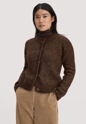 Frau trägt eine braune Strickjacke über einem dunkelbraunen Rollkragenpullover mit beigen Cordhosen, Hände in den Taschen, neutraler Hintergrund.