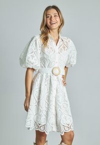 Biała sukienka z koronką, z bufiastymi rękawami, zapinana na guziki, z wąską talią i teksturowanym paskiem oraz falbaniastym dołem.