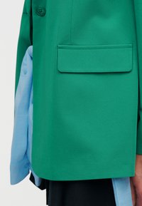 Gros plan sur un blazer vert à poche à rabat porté sur une chemise bleu clair et une jupe sombre, mettant en valeur les détails des manches et des boutons.