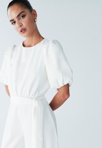 Witte jumpsuit met opgeblazen mouwen, ceintuuraan detail in de taille, gladde textuur en een ronde halslijn. Eenvoudig en elegant ontwerp.