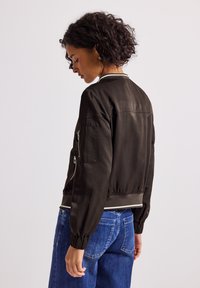 Femme aux cheveux bouclés portant une veste bomber foncée avec une poche zippée sur la manche et un jean bleu, de dos contre un fond uni.