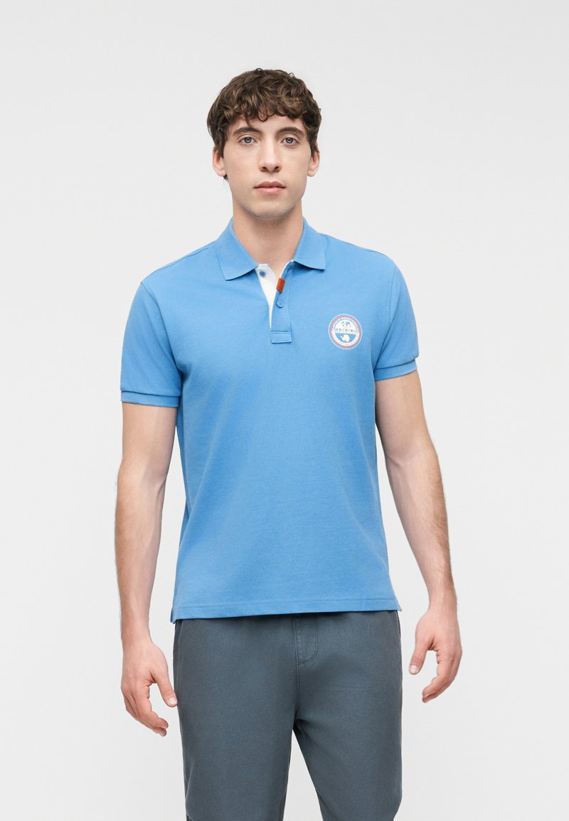 Άνδρας που φοράει ανοιχτό μπλε πόλο πουκάμισο με στρογγυλό λογότυπο στο αριστερό στήθος και σκούρο γκρι παντελόνι, στέκεται μπροστά σε απλό φόντο.