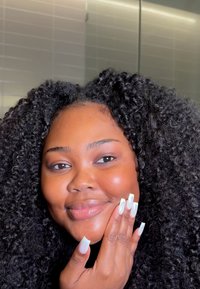 Svart kvinna med texturerat, voluminöst lockigt hår, leende och vilande sin hand på kinden, med långa, vitlackade naglar.