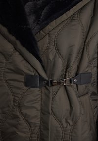 Oliwkowy pikowany płaszcz z miękkim czarnym futerkiem przy kołnierzu, z przodem zamykanym na ciemny skórzany pasek oraz srebrnymi elementami ozdobnymi.