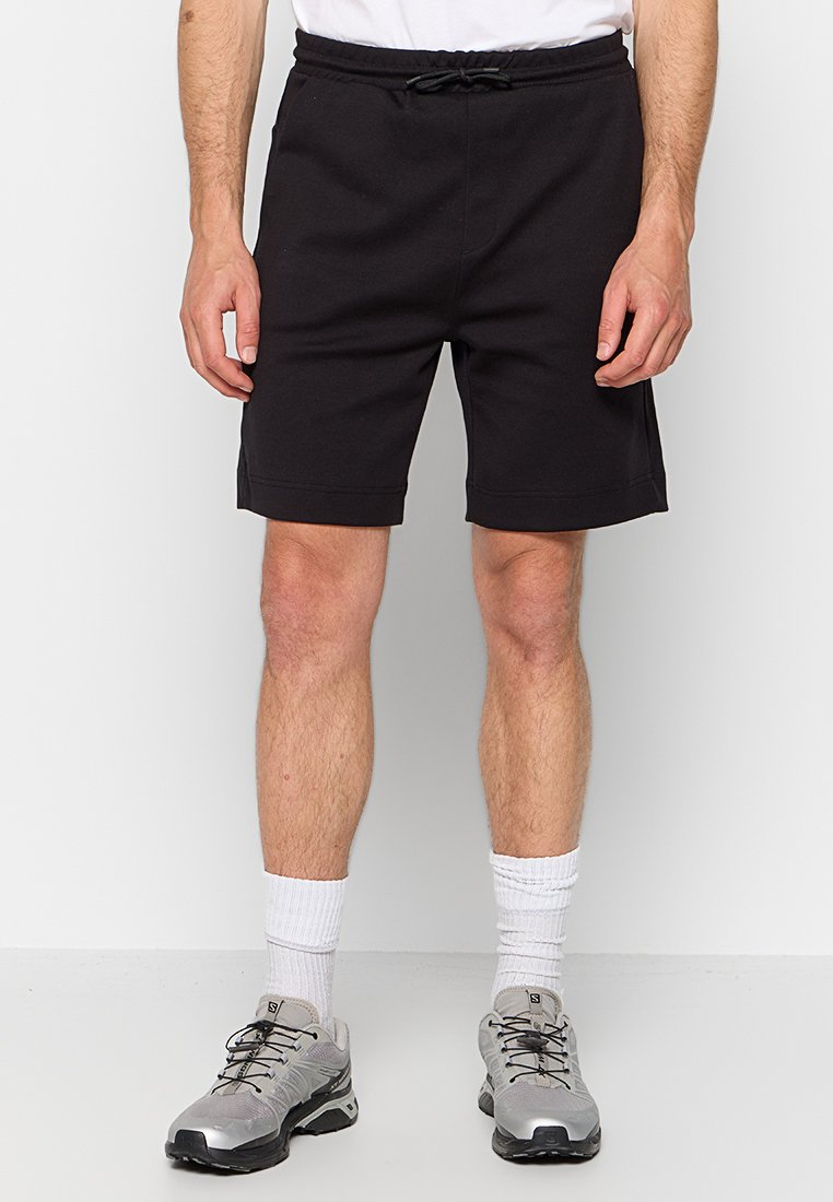 Czarne bawełniane szorty z elastycznym paskiem i sznurkiem, o prostym kroju i długości do kolan. Połączone z szarymi butami sportowymi.