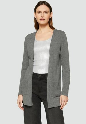 Frau trägt eine hellgraue, offene Strickjacke mit Taschen vorne über einem silbernen, ärmellosen Oberteil und schwarzen Hosen vor einem einfarbigen Hintergrund.