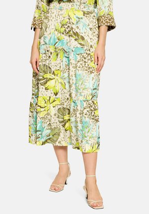 Femme portant une jupe midi fleurie avec des feuilles vertes, bleues et jaunes et des sandales beiges à talons à brides sur fond blanc.