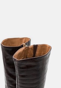Bottes en cuir marron foncé, montantes jusqu'au genou, avec une texture crocodile, dotées d'un intérieur beige et d'une fermeture éclair en haut.