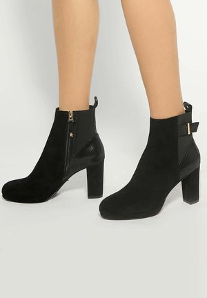 Jambes portant des bottines en daim noir avec talons blocs, fermetures éclair latérales et nœuds décoratifs sur un fond clair.