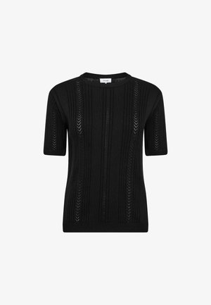 Vrouw staand met handen in zakken, draagt een zwart kortärmelig gebreid topje, wijde grijze broek en donkerbruine schoenen op een grijze vloer.