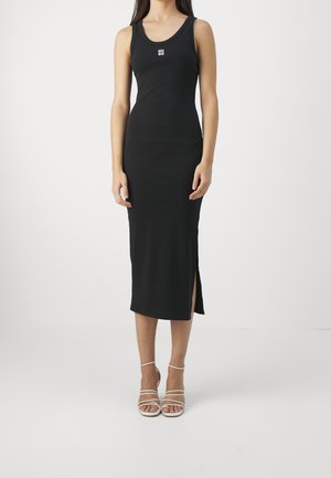 Kobieta ubrana w czarną, bezrękawnikową sukienkę midi z bocznym rozcięciem i białe, wiązane sandały na wysokim obcasie, stojąca na jednolitym tle.