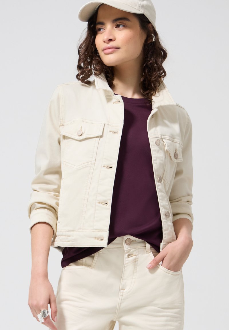 Femme aux cheveux bouclés portant une veste et un pantalon en jean crème, une chemise violet foncé et une casquette de baseball crème assortie, main dans la poche.
