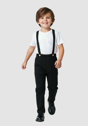 Czarne spodnie z szelkami, biała koszula z krótkim rękawem, błyszczące czarne buty i guziki przy spodniach. Chłopiec idzie z uśmiechem.