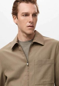 Giacca khaki con zip, colletto, tasca frontale e un maglione grigio texturizzato sotto. Tessuto liscio con una vestibilità strutturata.