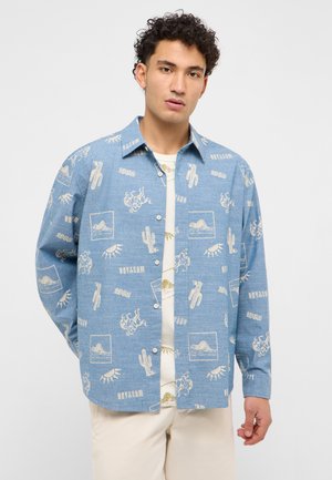 Junger Mann trägt ein blaues Hemd mit weißen Wüstenmotiven, darunter Kakteen, Pferde, Augen und Berge, über einem weißen T-Shirt.