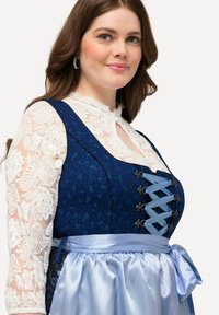 Vestido azul con estampado acompañado de una blusa blanca de encaje; cuenta con un corpiño con cintas y una falda de satén con un lazo atado en la cintura.