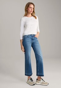 White long-sleeve top, blue wide-leg jeans with a frayed hem, and beige sneakers with black accents. Relaxed fit, casual style.