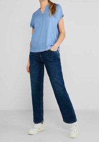 Světle modrý krátký rukáv top s výstřihem do V, v kombinaci s tmavě modrými rovnými džíny a bílými teniskami, umístěný na neutrálním pozadí.