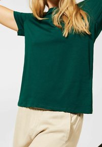 Grön bomullströja med korta ärmar och avslappnad passform, kombinerad med ljusbeiga byxor. Enkel design, slät textur, klassisk form.